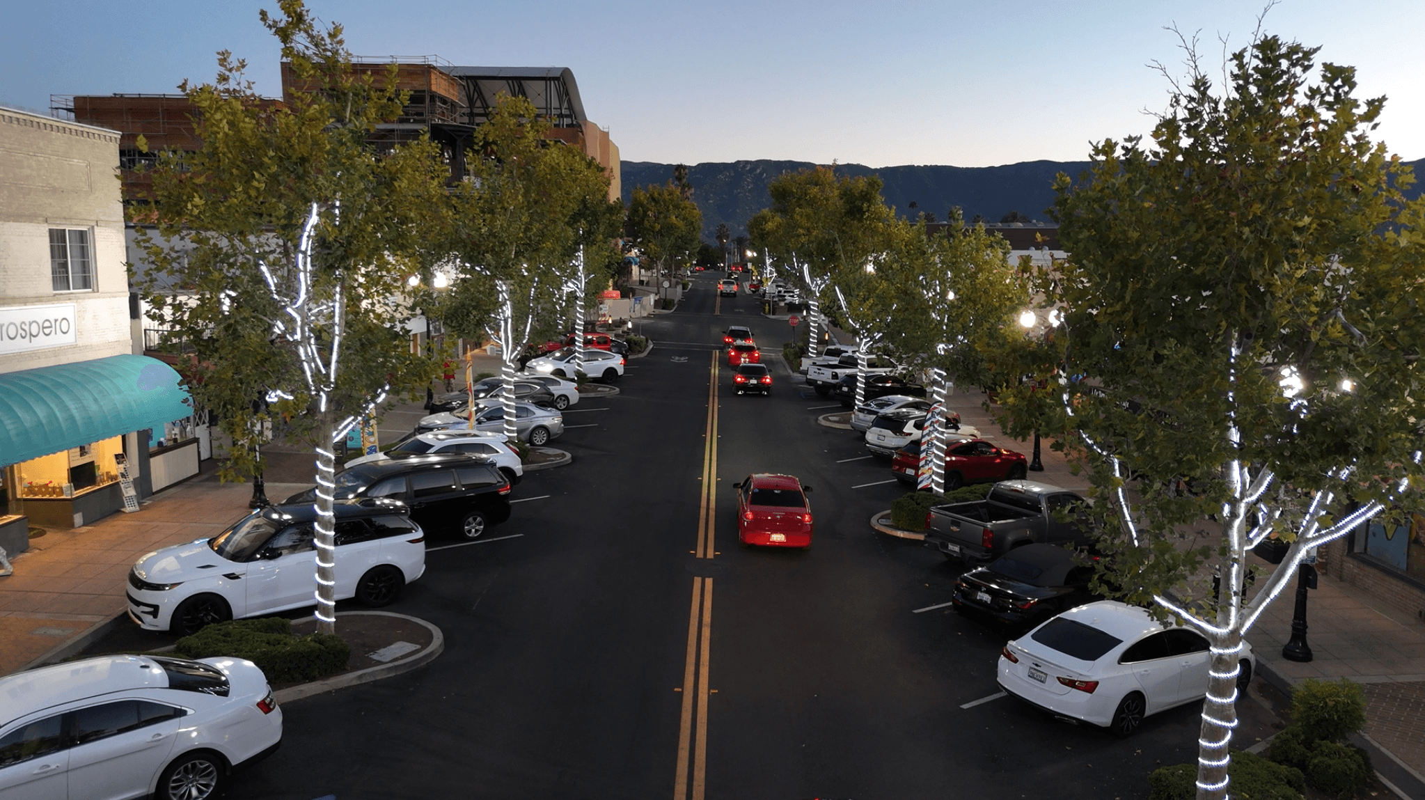 Evening street view with trees wrapped in white lights, parked cars, and distant mountains.